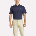 A man in a navy Reebok Men's BDC Signature Polo Shirt with RBK-DRY fabric, white chest logo, beige pants, and a striped belt stands against a plain white background.