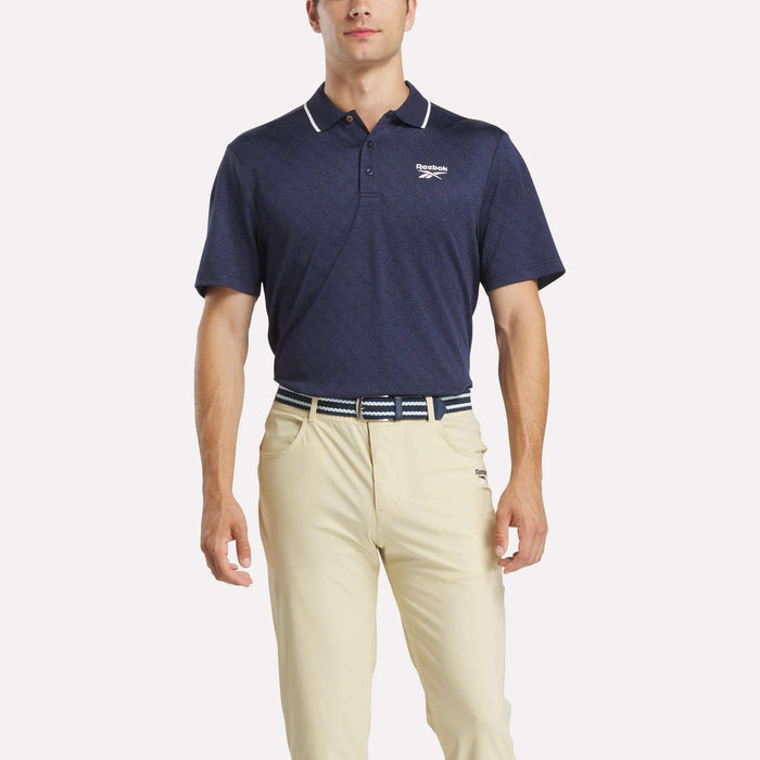 A man in a navy Reebok Men's BDC Signature Polo Shirt with RBK-DRY fabric, white chest logo, beige pants, and a striped belt stands against a plain white background.