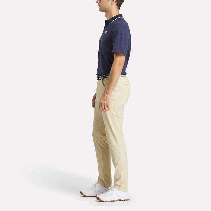 A man in profile wears the Reebok Men's BDC Signature Polo Shirt, beige pants, a white belt, and white shoes, standing against a plain white background.