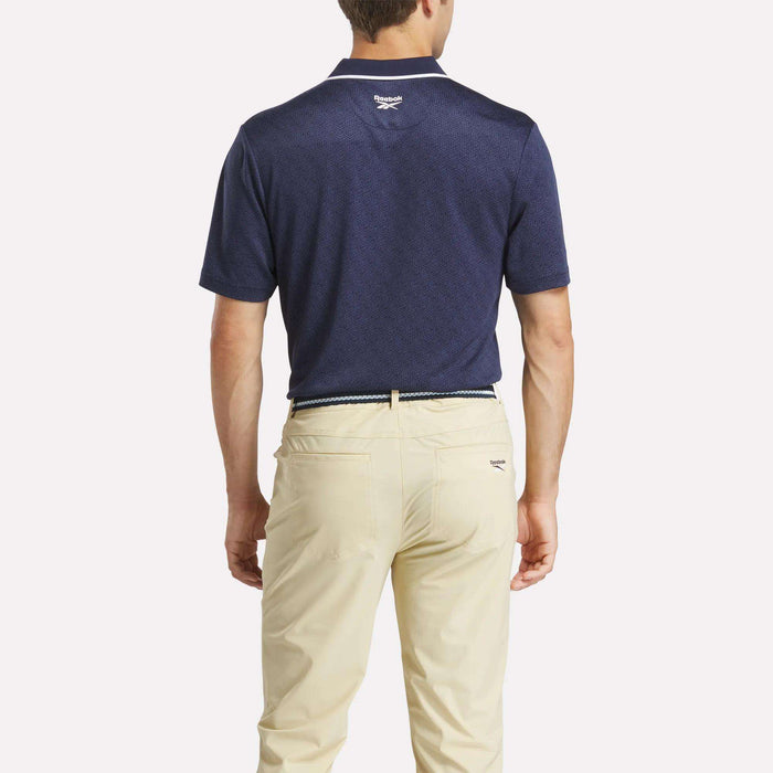 A man, seen from behind, wears a Reebok Men's BDC Signature Polo Shirt in navy blue RBK-DRY UPF fabric, tucked into light beige pants. A small white logo appears under the collar.