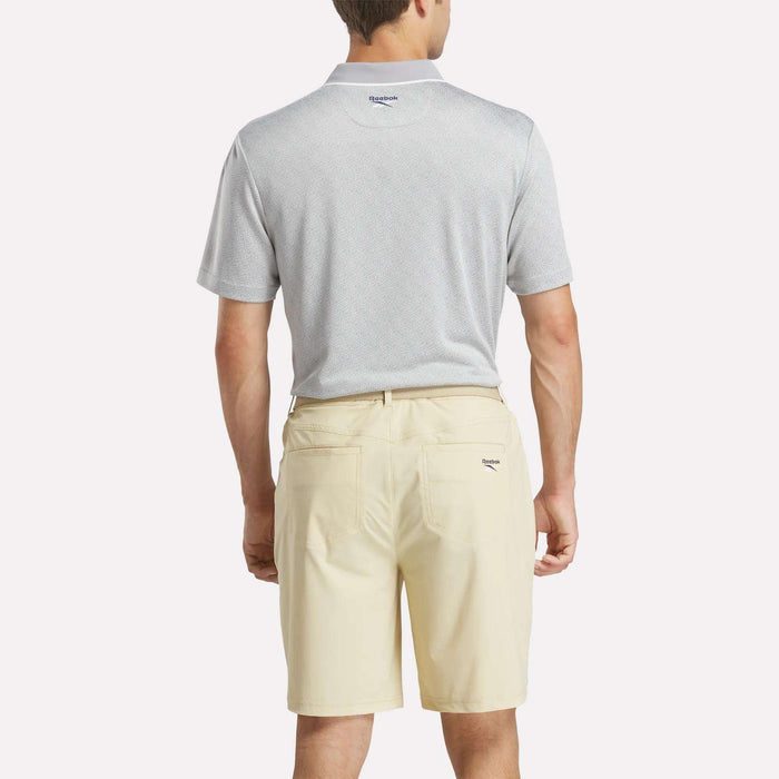 A man is seen from the back wearing a light gray Reebok Men's BDC Signature Polo Shirt and cream-colored shorts, standing against a simple, light background.