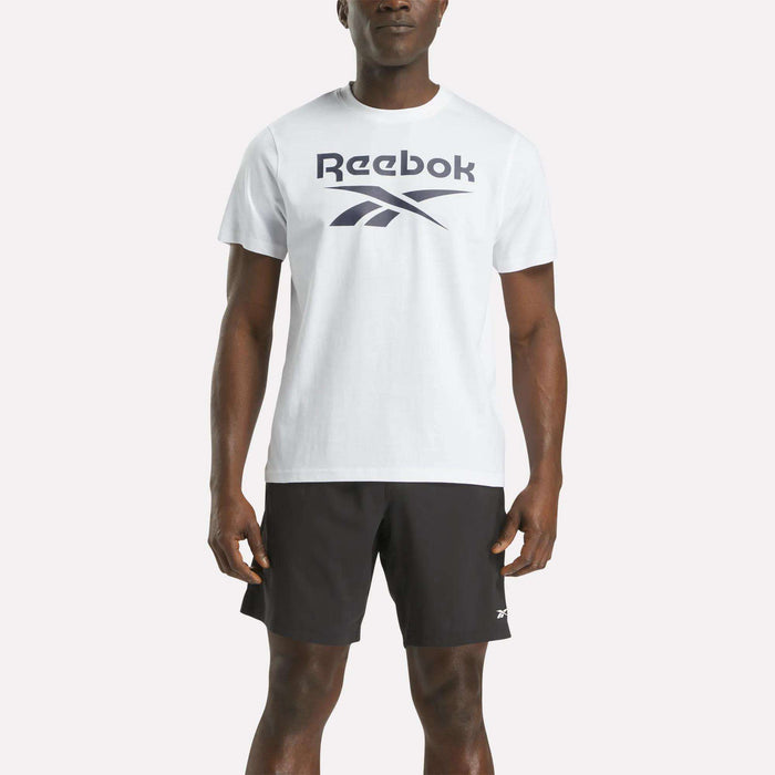 A person stands against a plain light background wearing a Reebok Men's Big Logo Crewneck Short Sleeve Tee in white and black athletic shorts.