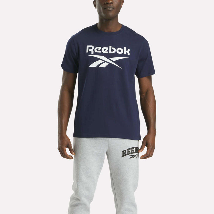 A person faces forward wearing the Reebok Men's Big Logo Crewneck Short Sleeve Tee in navy with a large white logo, paired with light gray sweatpants featuring a small logo near the left pocket.