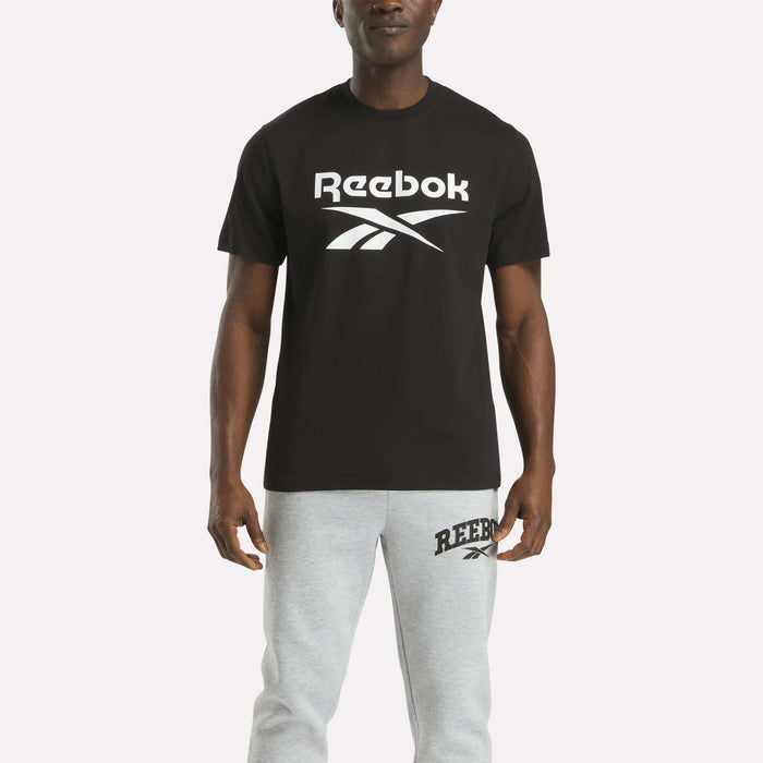 A person faces forward against a plain white background, wearing the Reebok Men's Big Logo Crewneck Short Sleeve Tee in black with a large white logo and light gray Reebok sweatpants.