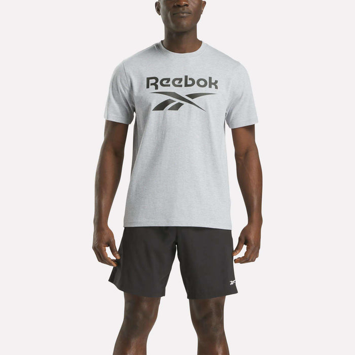 A man wearing a Reebok Men's Big Logo Crewneck Short Sleeve Tee in light gray with a black logo and black athletic shorts stands facing forward against a plain white background.