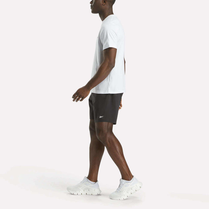 A man wears a Reebok Men's Big Logo Crewneck Short Sleeve Tee and black athletic shorts with white sneakers featuring a subtle Reebok logo, walking forward against a plain white background with his left arm slightly extended.