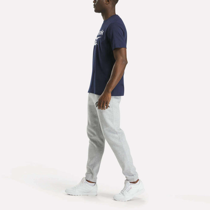 A man stands sideways against a plain white background wearing a Reebok Men's Big Logo Crewneck Short Sleeve Tee in navy blue, light gray jogger pants, and white sneakers.