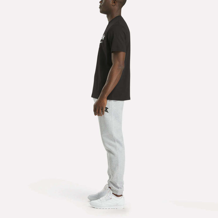 A man stands in profile against a plain white background, wearing Reebok's Men's Big Logo Crewneck Short Sleeve Tee, paired with light gray jogger pants and white sneakers. The tee features a bold Reebok logo on the chest.