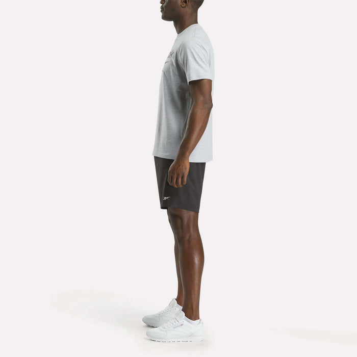 A man in side profile wears the Reebok Men's Big Logo Crewneck Short Sleeve Tee in light grey, paired with black athletic shorts and white sneakers, set against a plain white background.