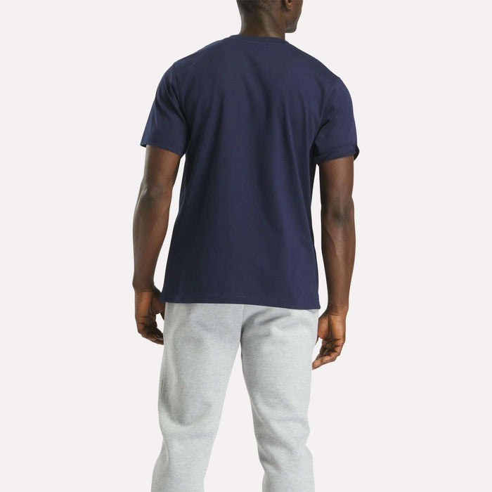 A person faces away from the camera, wearing a Reebok Men's Big Logo Crewneck Short Sleeve Tee in navy blue and light gray sweatpants, set against a plain light background.
