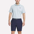 A man stands against a plain white background wearing a light blue Reebok Men's Bonded Pique Polo Shirt with a small left chest logo, navy shorts, and a patterned belt.