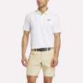 A man in a white Reebok Men's Bonded Pique Polo Shirt, beige shorts, and a black belt stands against a plain white background with his head slightly turned and arms relaxed at his sides.