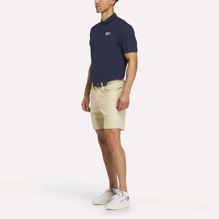 A man stands against a plain white background wearing a Reebok Men's Bonded Pique Polo Shirt in navy blue, beige shorts, a black belt, and white sneakers.