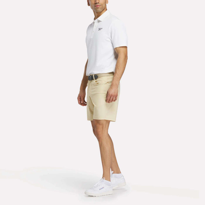 A man faces slightly left, wearing a white Reebok Men's Bonded Pique Polo Shirt, beige shorts, a black belt, and white sneakers against a plain white background.