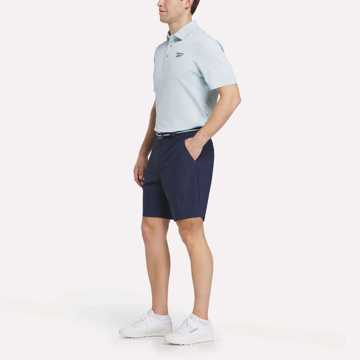 A man stands with hands in pockets, wearing a light blue Reebok Men's Bonded Pique Polo Shirt, navy shorts, a belt, and white sneakers against a plain white background.