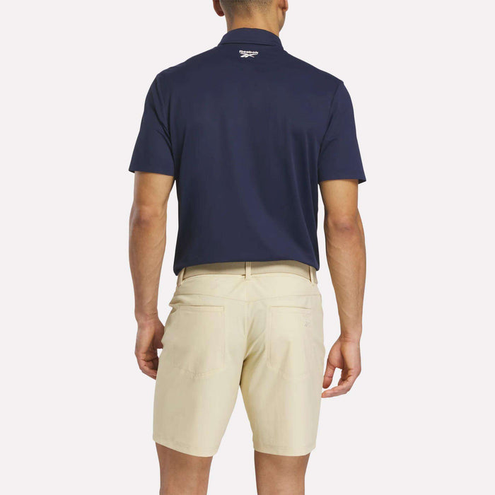 A man faces away from the camera, wearing a navy Reebok Men's Bonded Pique Polo Shirt and beige shorts. His arms rest at his sides against a plain, light background.