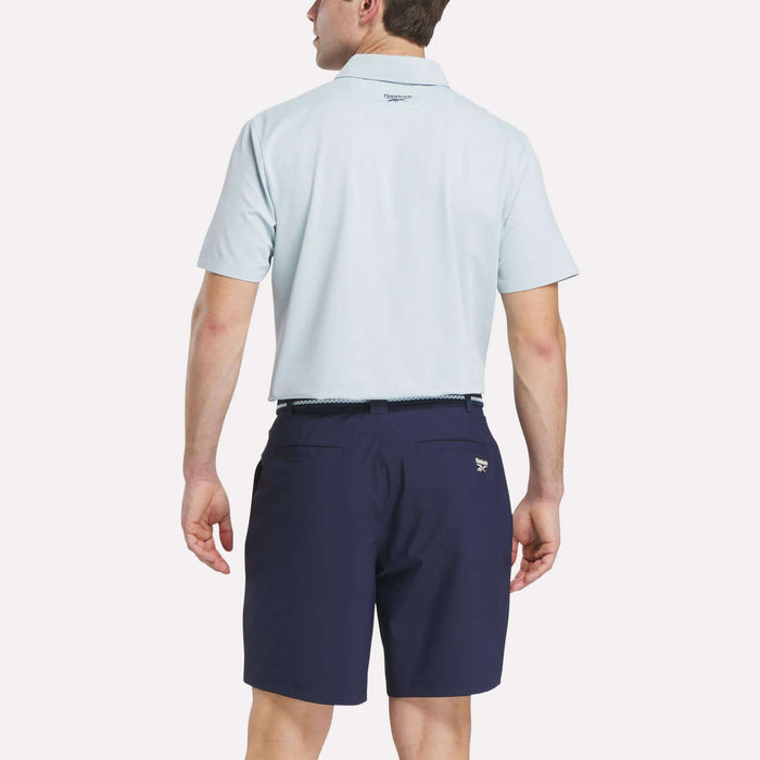 A man faces away from the camera, wearing a light blue Reebok Men's Bonded Pique Polo Shirt tucked into navy blue shorts with a belt against a plain, light background.