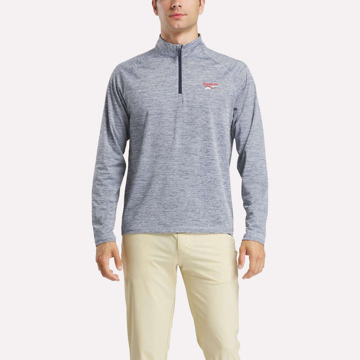 A man stands facing forward, arms at his sides, wearing a Reebok Men's Cationic Wicking Quarter-Zip Sweatshirt in light blue and beige pants against a plain white background.