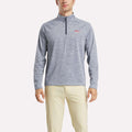 A man stands facing forward, arms at his sides, wearing a Reebok Men's Cationic Wicking Quarter-Zip Sweatshirt in light blue and beige pants against a plain white background.