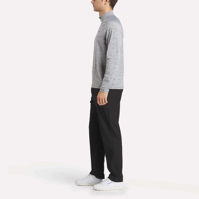 A man stands in profile wearing a Reebok Men's Cationic Wicking Quarter-Zip Sweatshirt, black pants, and white sneakers against a plain white background.