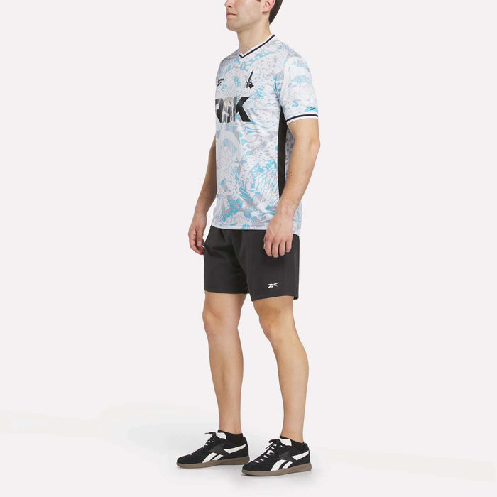 A man stands wearing the Men's Charlton Away Jersey by Reebok, paired with black athletic shorts and black sneakers, set against a plain white background.