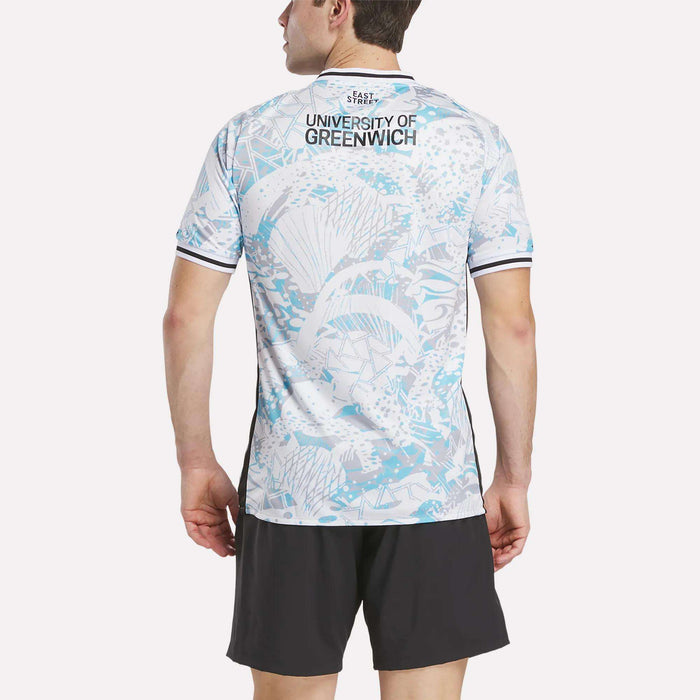 A man faces away wearing the Reebok Men's Charlton Away Jersey, featuring an aquatic blue and white heritage-inspired design with "University of Greenwich" on the back, paired with black athletic shorts.