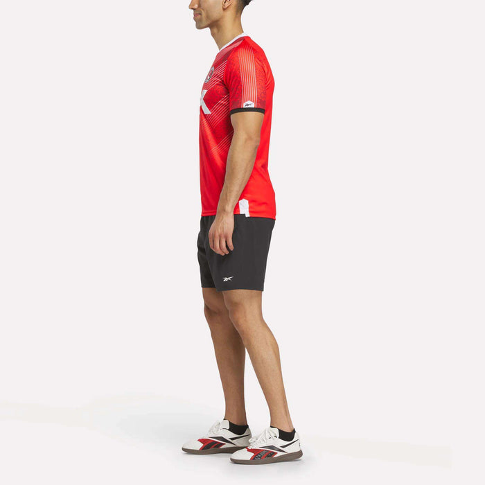 A man stands sideways in a Reebok Men's Charlton Home Jersey with club badge, black athletic shorts, and white sneakers featuring red and black details against a plain white background.