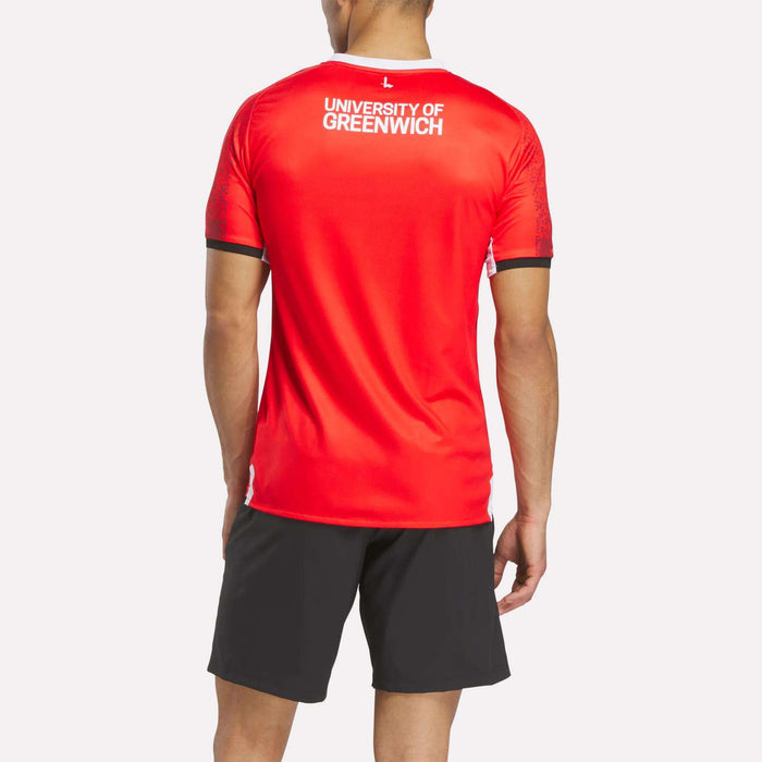 A person faces away, wearing the Reebok Men's Charlton Home Jersey in bright red with club badge details and black athletic shorts, set against a plain white background.