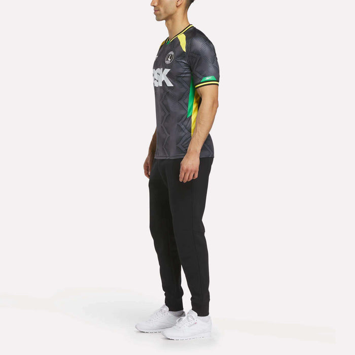 A man stands in profile against a white background, wearing the Reebok Men's Charlton Third Jersey with black pants and white sneakers. The shirt features white lettering plus green and yellow accents inspired by the team's third kit.