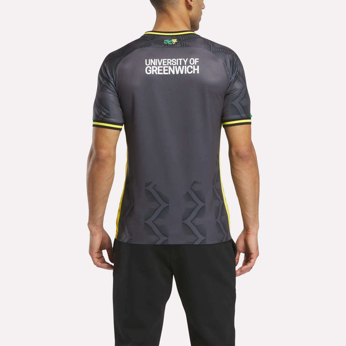 A man faces away from the camera wearing the Reebok Men's Charlton Third Jersey, a dark athletic shirt with yellow accents, University of Greenwich on the upper back, and lightweight fabric. He pairs it with black pants.