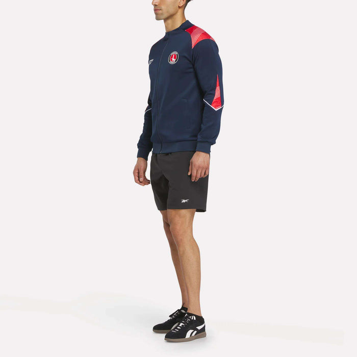 A man in a Reebok Men's Charlton Training Jacket paired with black shorts and sneakers stands facing left against a plain white background.