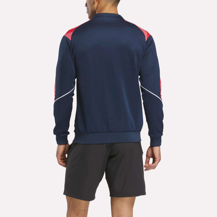 A man stands with his back to the camera in a Reebok Men's Charlton Training Jacket featuring red and white shoulder and sleeve accents, made from polyester doubleknit, paired with black shorts against a plain background.