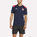 A man wears a navy blue Reebok Men's Charlton Training T-Shirt with red and white shoulder accents, paired with black shorts, standing against a plain light background.