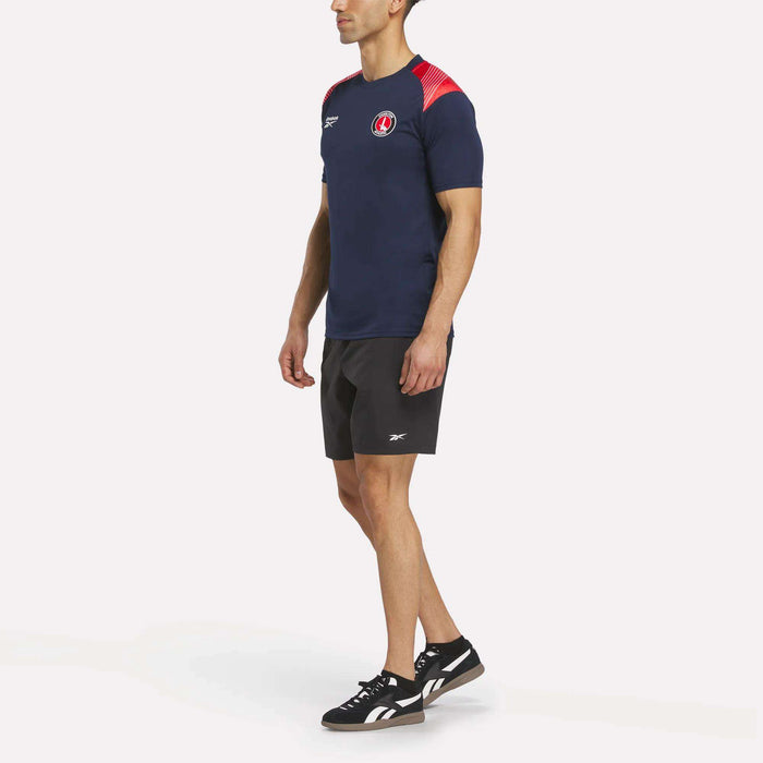 A man faces slightly left, wearing the Reebok Men's Charlton Training T-Shirt in navy blue with red accents, paired with black shorts and black sneakers with white soles. The shirt is made from breathable polyester for comfort.