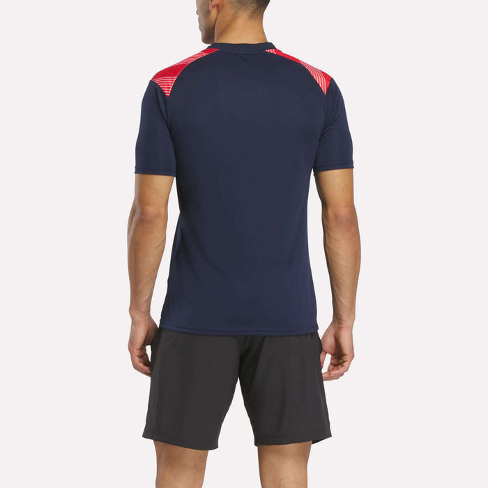 A man seen from behind wears a Reebok Men's Charlton Training T-Shirt in navy with red shoulder accents and black shorts, made from breathable polyester, standing against a plain light background.