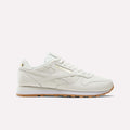 A Men's Classic Leather Shoe by Reebok in Chalk/Black/Ftwrwhite, featuring a gold logo accent, white laces, and a light brown rubber sole with an EVA midsole for lightweight cushioning, is displayed in a side profile against a plain light background.
