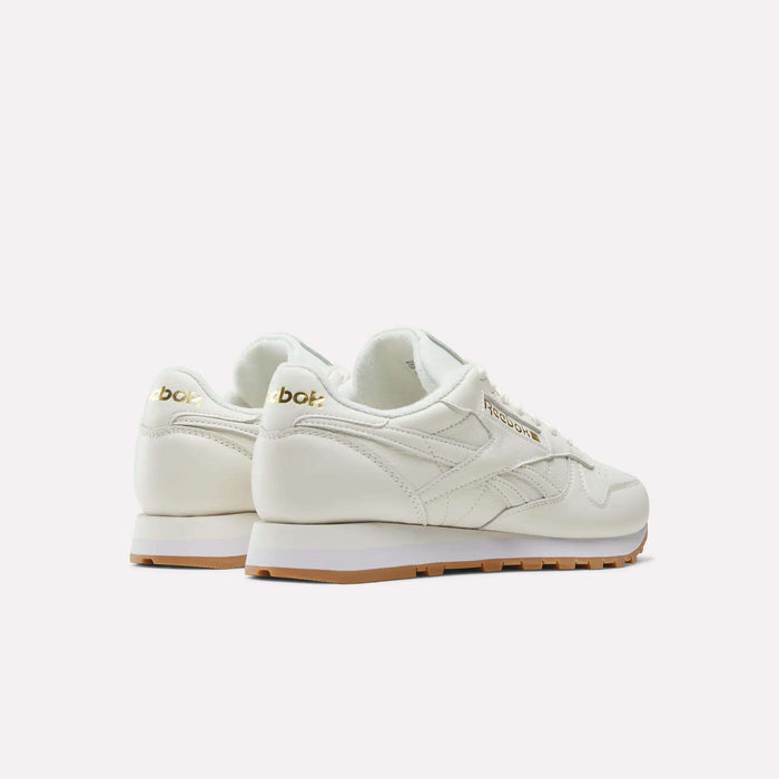 A pair of Men's Classic Leather Shoes by Reebok in off-white, featuring gold logo accents, gum rubber soles, and a cushioned EVA midsole, displayed from rear and side views against a plain white background.