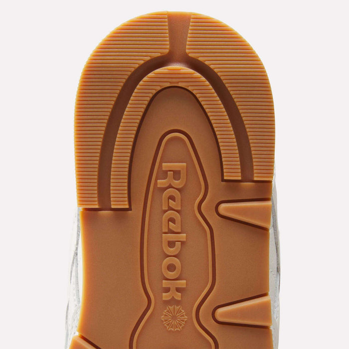Close-up of the sole of the Reebok Men's Classic Leather Shoes, featuring the Reebok logo and a brown tread pattern with textured grooves, supported by an EVA midsole, displayed against a white background.