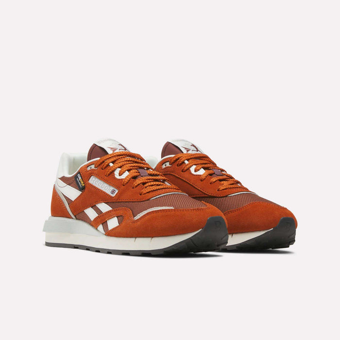 A pair of Reebok Men’s Classic Nylon 89 Shoes in orange and white, crafted from mixed materials with white soles, orange laces, black accents, and a comfortable EVA midsole, are positioned side by side on a plain white background.