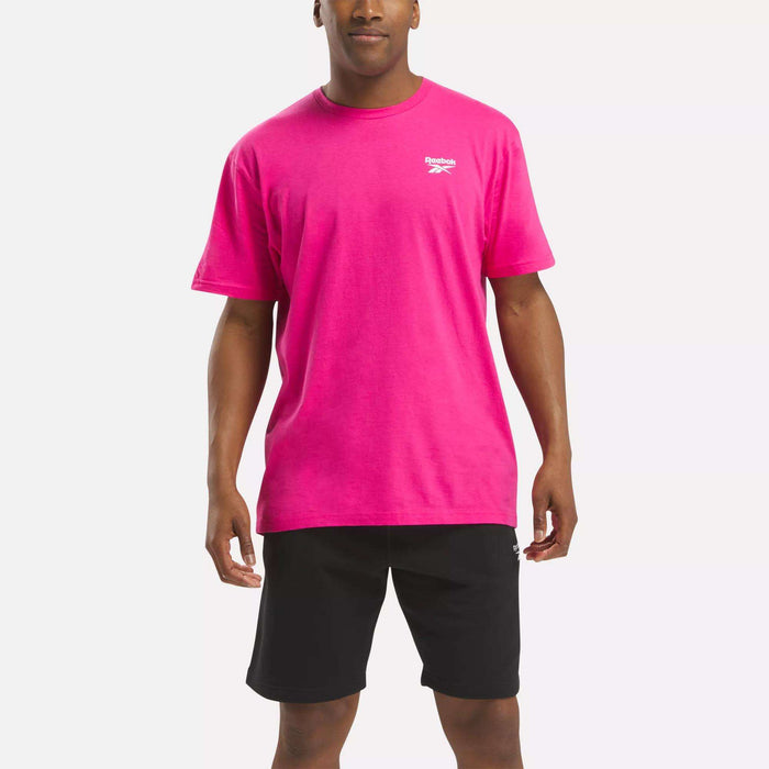 A man stands facing forward against a plain white background, wearing a bright pink Reebok Men's Classic Small Logo T-Shirt made of soft cotton and black shorts.