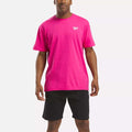 A man stands facing forward against a plain white background, wearing a bright pink Reebok Men's Classic Small Logo T-Shirt made of soft cotton and black shorts.