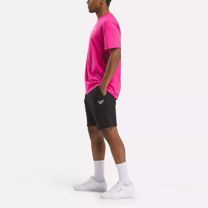 A person stands sideways with hands in pockets, wearing a bright pink Reebok Men's Classic Small Logo T-Shirt, black shorts, white crew socks, and white sneakers against a plain white background.