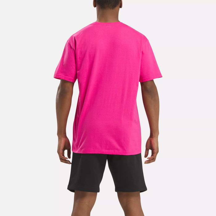 A person is standing with their back to the camera, wearing a bright pink Reebok Men's Classic Small Logo T-Shirt and black shorts against a plain light background.