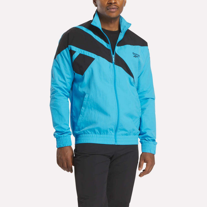 A man models the Reebok Men's Classics Track Top in bright blue with black geometric chest and shoulder patterns, paired with black pants, standing against a plain light background to showcase casual comfort.