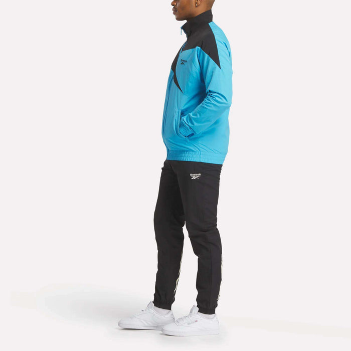 A person stands sideways with hands in pockets, wearing the Reebok Men's Classics Track Top in blue and black with white sneakers, effortlessly embodying casual comfort against a plain white background.
