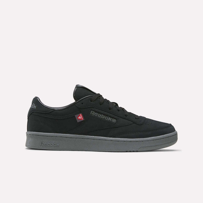 A pair of Reebok Men’s Club C 85 Shoes in Black/Grey 4, featuring a gray sole, black laces, and a subtle red Reebok logo on the side, is displayed against a plain white background. These classic sneakers offer a sleek design paired with everyday comfort.