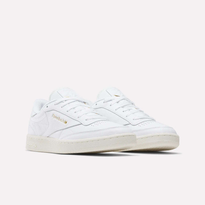 The Reebok Men's Club C 85 Shoes feature white premium garment leather, off-white soles, gold Reebok branding on the side, and a classic low-top silhouette. Displayed against a plain white background.