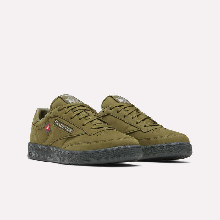 A pair of Men’s Club C 85 Shoes by Reebok in olive green, featuring black soles, signature Reebok branding, and a small red tag on the side, displayed against a plain white background.