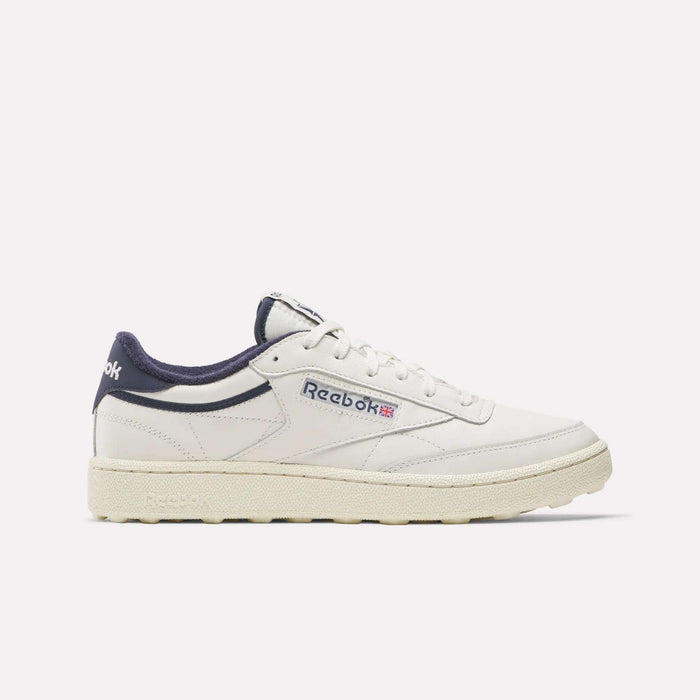 The Reebok Men's Club C Golf Shoes are white with navy blue accents, a beige spikeless outsole, and feature the Reebok logo with a small Union Jack on the side, displayed in a side profile on a plain white background.