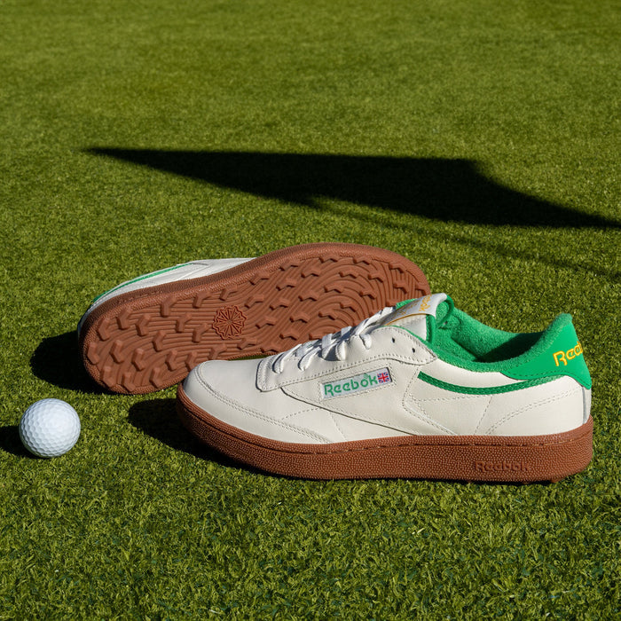 Two Reebok Men's Club C Golf Shoes with white uppers, green accents, and spikeless outsoles rest on a green grass surface beside a white golf ball.